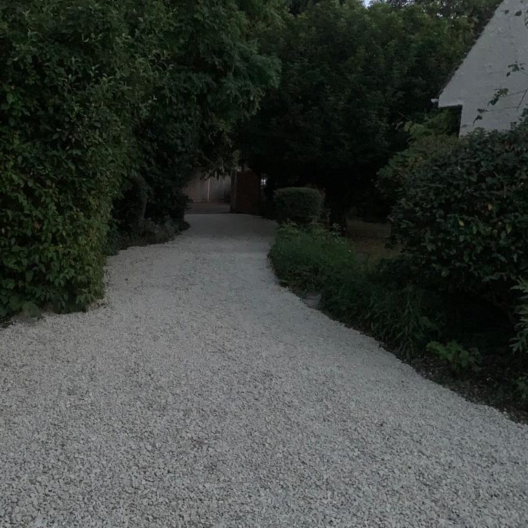 Gravel driveway leading through lush greenery towards a house in the background.