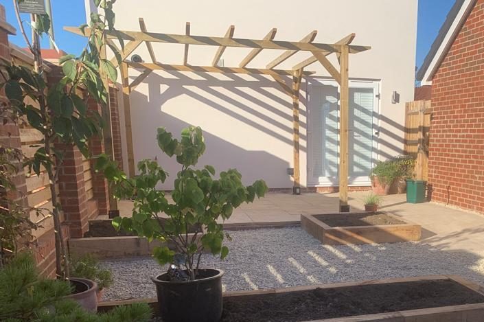A wooden pergola casts shadows over a gravel patio, featuring potted plants.