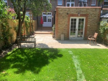 A sunny garden with a lawn, seating areas, and a brick wall building in the background.
