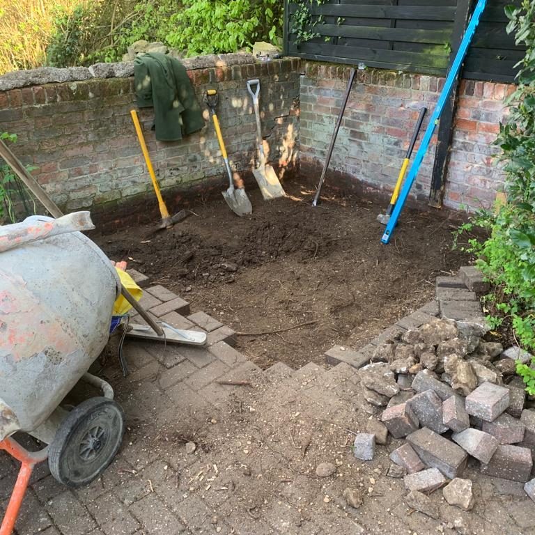 A dig site with tools and fresh soil surrounded by bricks and a wheelbarrow.