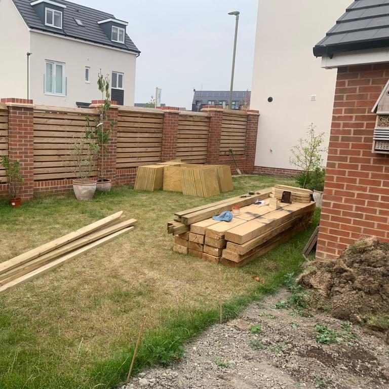 A garden area with building materials, including planks and a wooden structure, near a house.