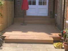 Patio area with wooden steps, planters, and a red parasol beside a beige wall.