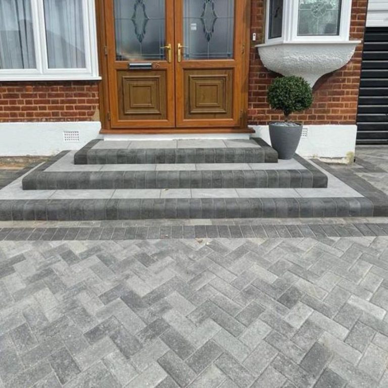 Grey herringbone-patterned paving leading to wooden front doors, flanked by decorative plants.