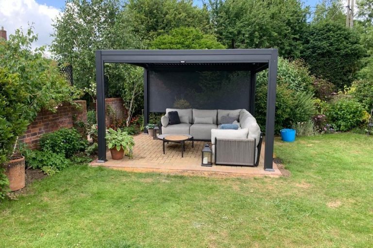 Garden seating area under a black pergola with a grey sofa and table surrounded by greenery.