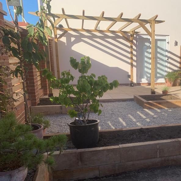 Garden with wooden pergola, gravel pathway, and potted plants in sunlight.