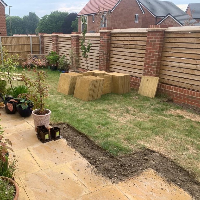 Garden scene with paving stones, plants, and hay bales against a wooden fence.